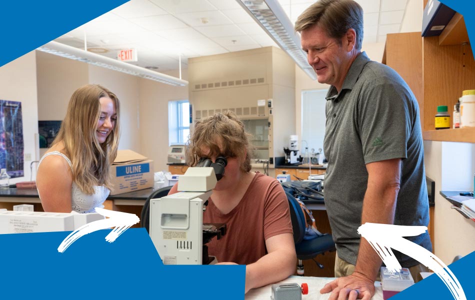 Two students and an instructor work together in a science laboratory. One student looks through a microscope while another stands nearby, and the instructor observes the activity. Laboratory equipment, workstations, and storage cabinets are visible in the background. Blue graphic accents frame the image along the top and bottom edges.