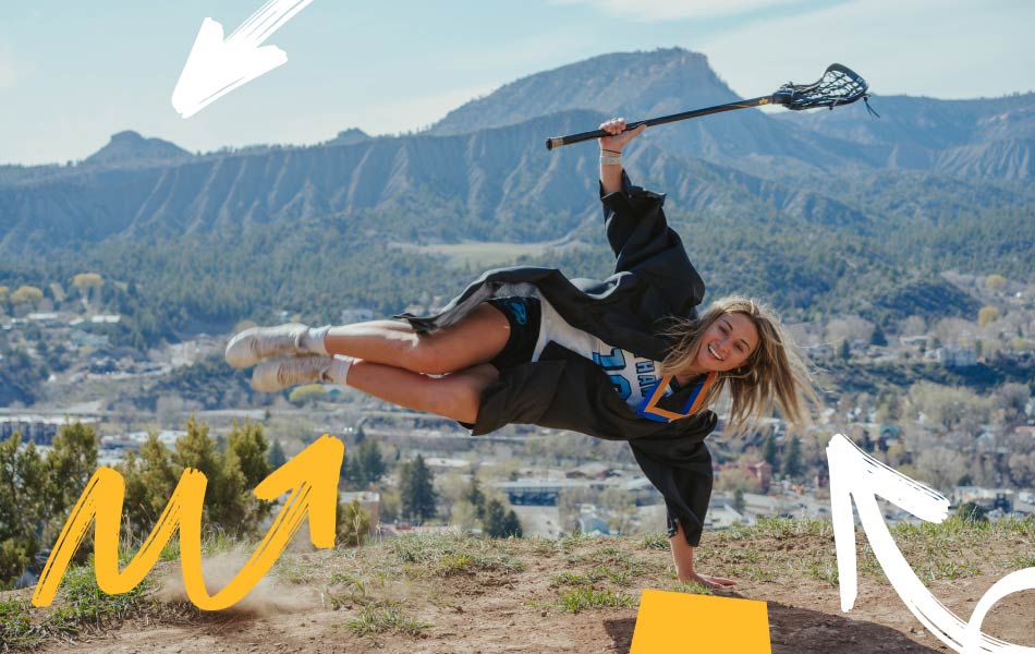Graduate wearing academic regalia and a medal performing a one‑handed pose while holding a lacrosse stick on a hillside overlook, with Durango and surrounding mountains visible in the background.