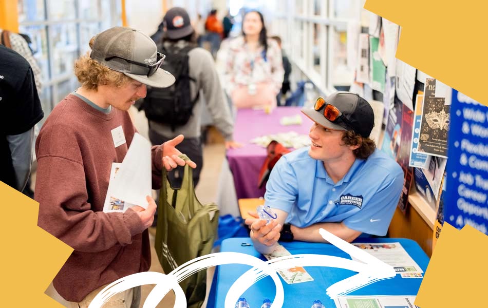 Student speaking with an organization representative at an indoor campus information fair, with brochures, displays, and attendees visible along a hallway lined with tables.