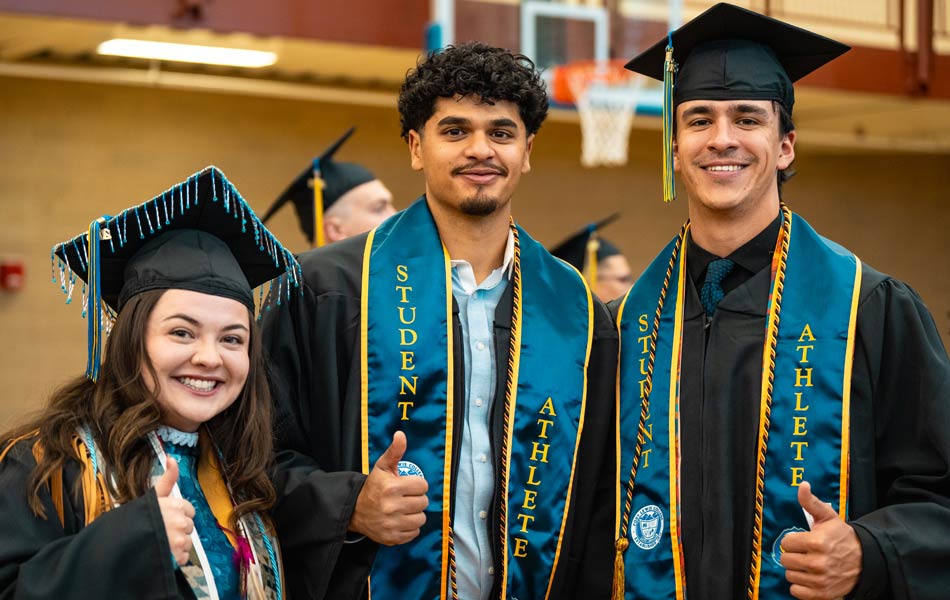 Three graduates wearing caps and gowns stand together indoors, smiling and giving thumbs up, with student‑athlete stoles visible.