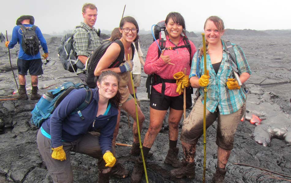 Group of students wearing backpacks and work gloves standing on a rugged lava field during an outdoor field study.