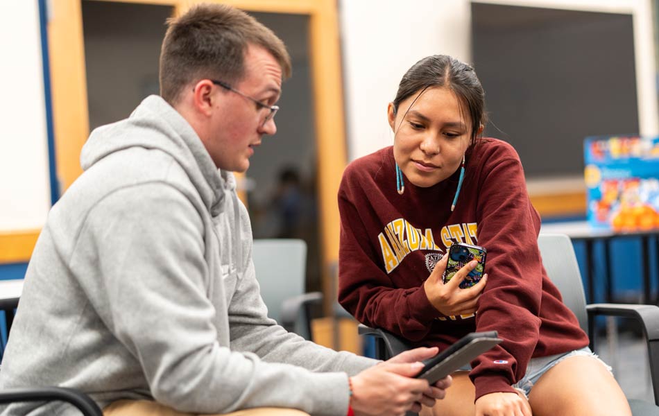 Two students seated in a classroom reviewing content on a tablet and smartphone together in a small‑group learning setting.