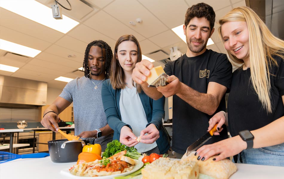 Four students working together in a teaching kitchen, preparing food at a counter with vegetables, bread, and cooking equipment, while one student sprinkles an ingredient onto a dish.
