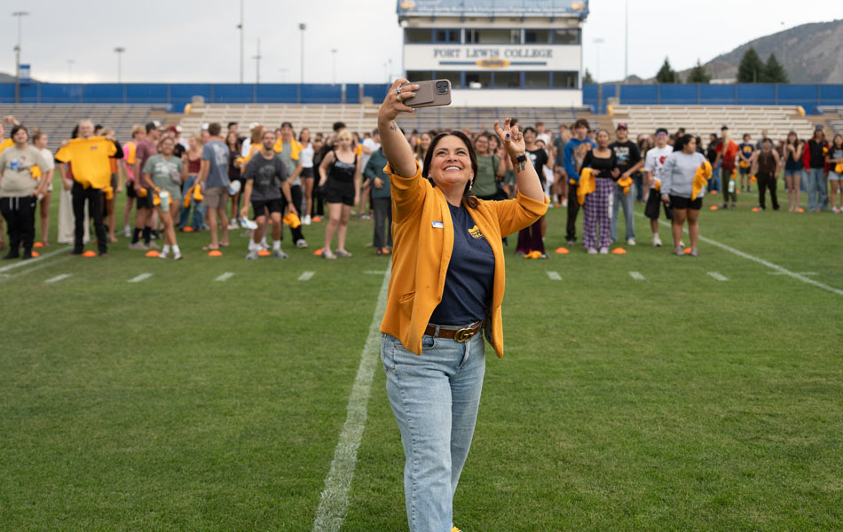 President Heather standing on a football field taking a selfie, with a large group gathered behind them and Fort Lewis College stadium seating in the background.