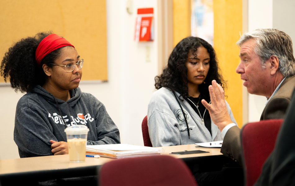 Faculty member seated at a table speaking with two students during a classroom discussion, with notebooks and learning materials visible.