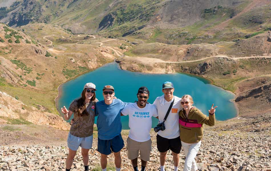Group of students standing on a rocky hillside overlooking a bright blue alpine lake, with rugged mountain terrain and winding trails in the background.