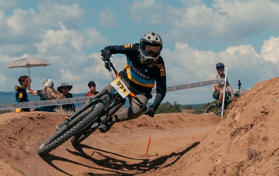 Mountain biker racing through a dirt course during a competition, leaning into a bermed turn with spectators and race tape visible in the background.