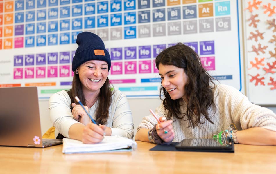 Two students seated at a table working together on coursework, with notebooks, pencils, a laptop, and a periodic table displayed on the wall behind them.