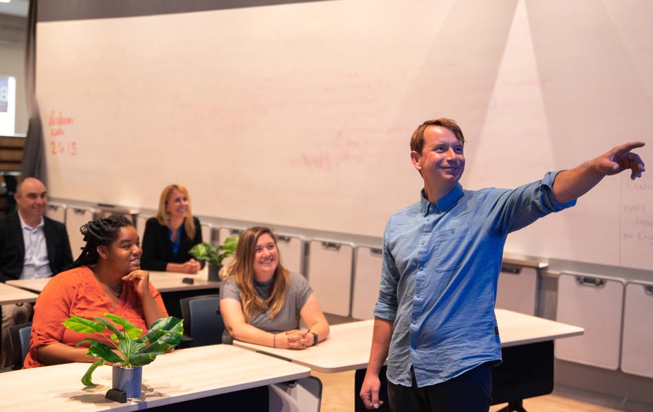 Instructor standing at the front of a classroom pointing toward a whiteboard, with several participants seated at tables engaged in a lesson.