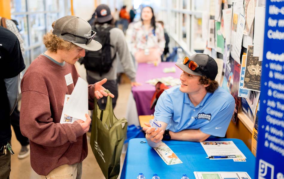 Two people talking at a table during a campus event, with informational materials, posters, and attendees visible in a hallway setting.