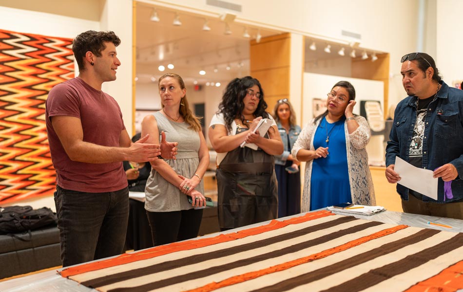 Small group gathered around a textile laid out on a table in a gallery setting, listening as one person gestures while others take notes, with woven artwork displayed on the walls behind them.
