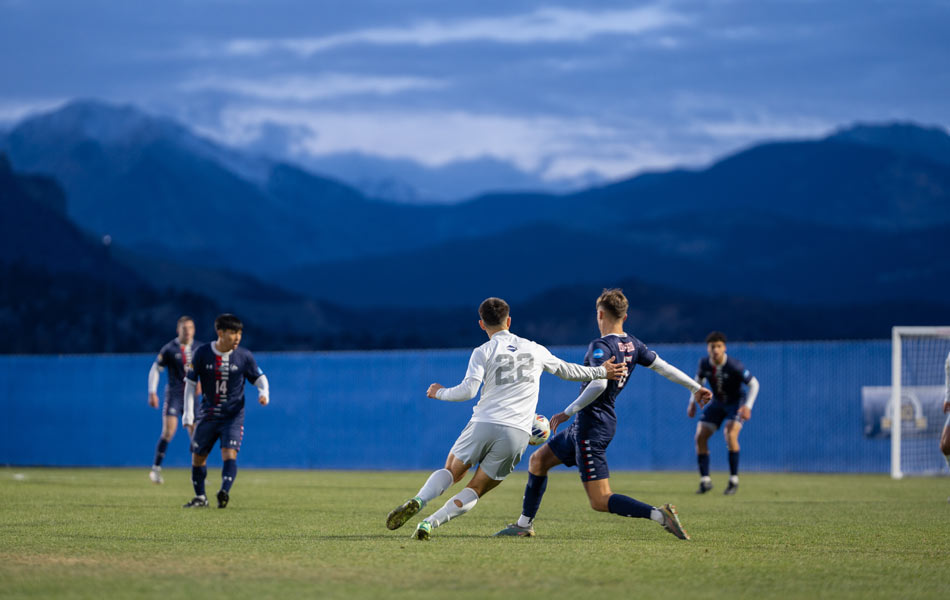 College soccer players competing on a field at dusk, with stadium lights on and mountain peaks visible beyond the far sideline.