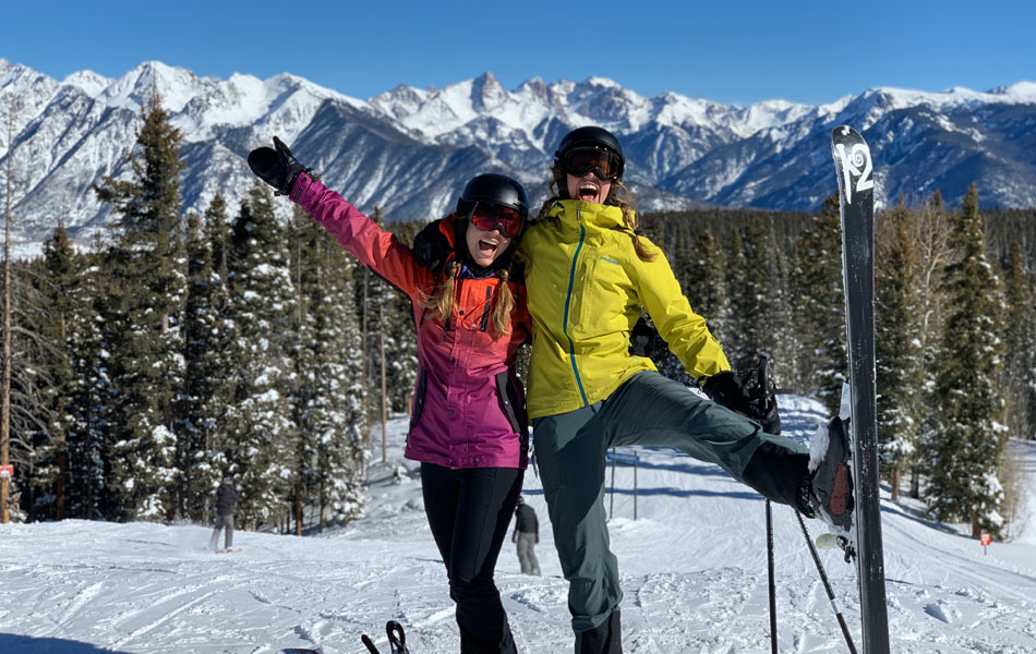 Two skiers posing on a snowy slope at Purgatory Resort, with skis raised and snow‑covered mountain peaks and evergreen trees in the background.